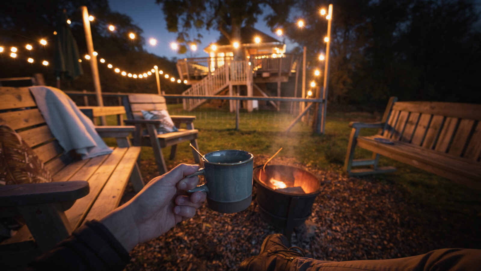 A person holds a mug near a fire pit, surrounded by wooden benches and twinkling string lights, with the cozy Trawscwm Treehouse nestled in the background.