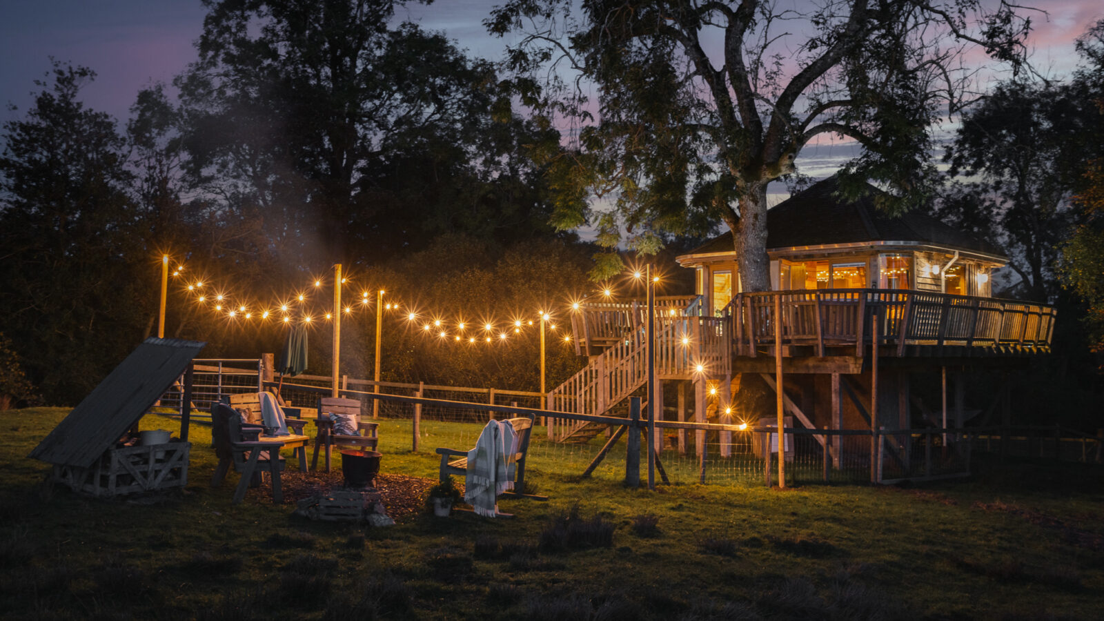 The Trawscwm Treehouse glows warmly at night, adorned with string lights and surrounded by inviting outdoor seating. A cozy fire pit area beneath the dark sky completes this enchanting scene.