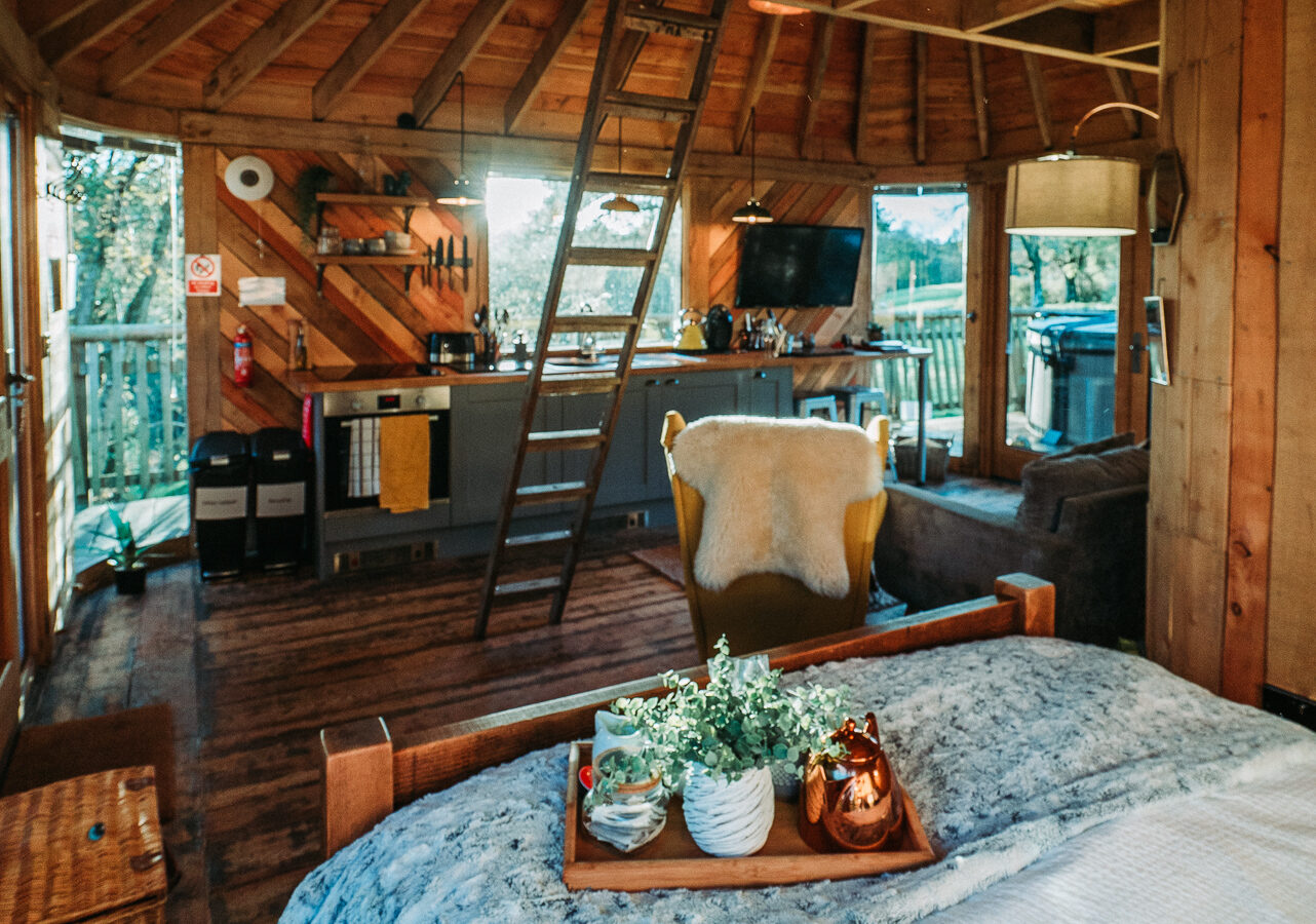 The interior of Trawscwm Treehouse features wooden beams, a comfy bed, a cozy chair, and a small kitchen area. A ladder leads to the loft in this rustic cabin atmosphere.