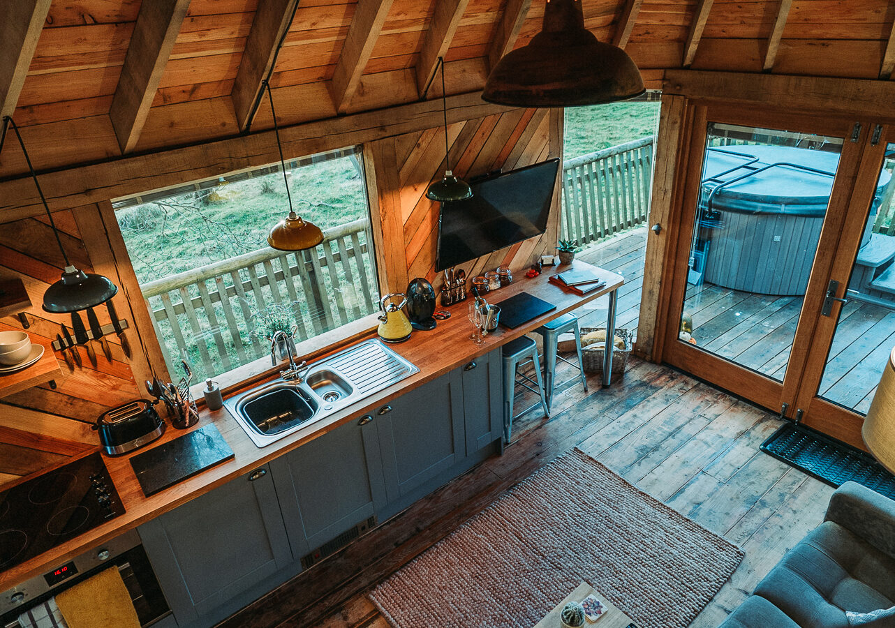 The Trawscwm Treehouse offers a cozy cabin interior with wooden walls, a quaint kitchen area, and a dining table, all complemented by large windows that provide a stunning view of the fenced yard.