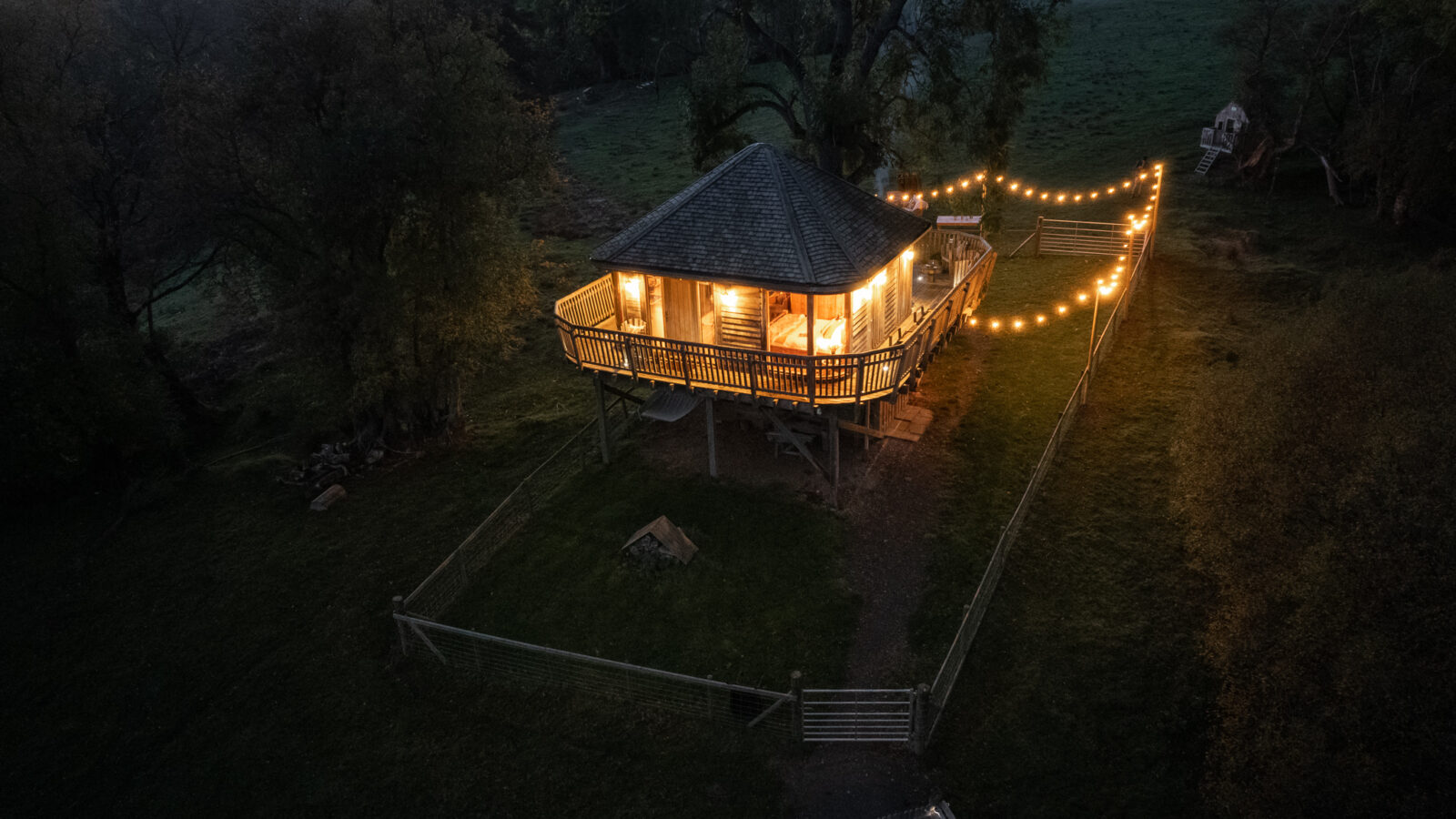 A wooden cabin with a lit porch and string lights in a grassy area surrounded by trees at dusk, offering a unique stay reminiscent of Trawscwm Treehouse.