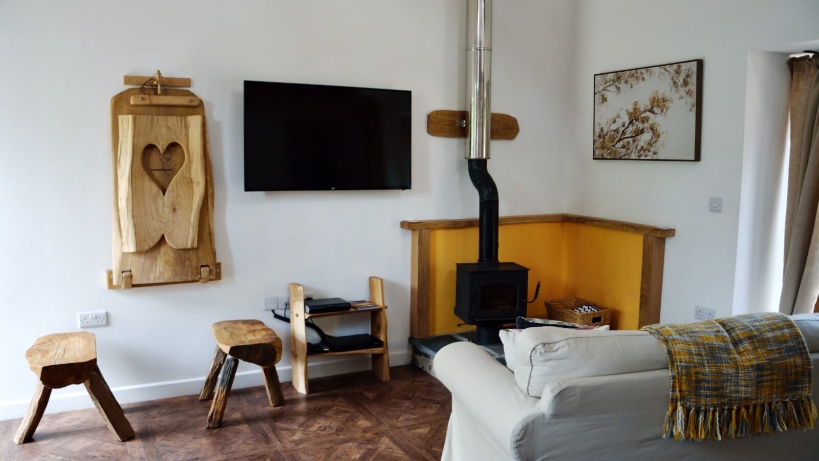 Explore the charming living room at Nant Awen Cottages, featuring a mounted TV, wooden stools, a wood-burning stove, and a cozy sofa draped with a blanket. Rustic decor elements add to the cottage's inviting atmosphere.