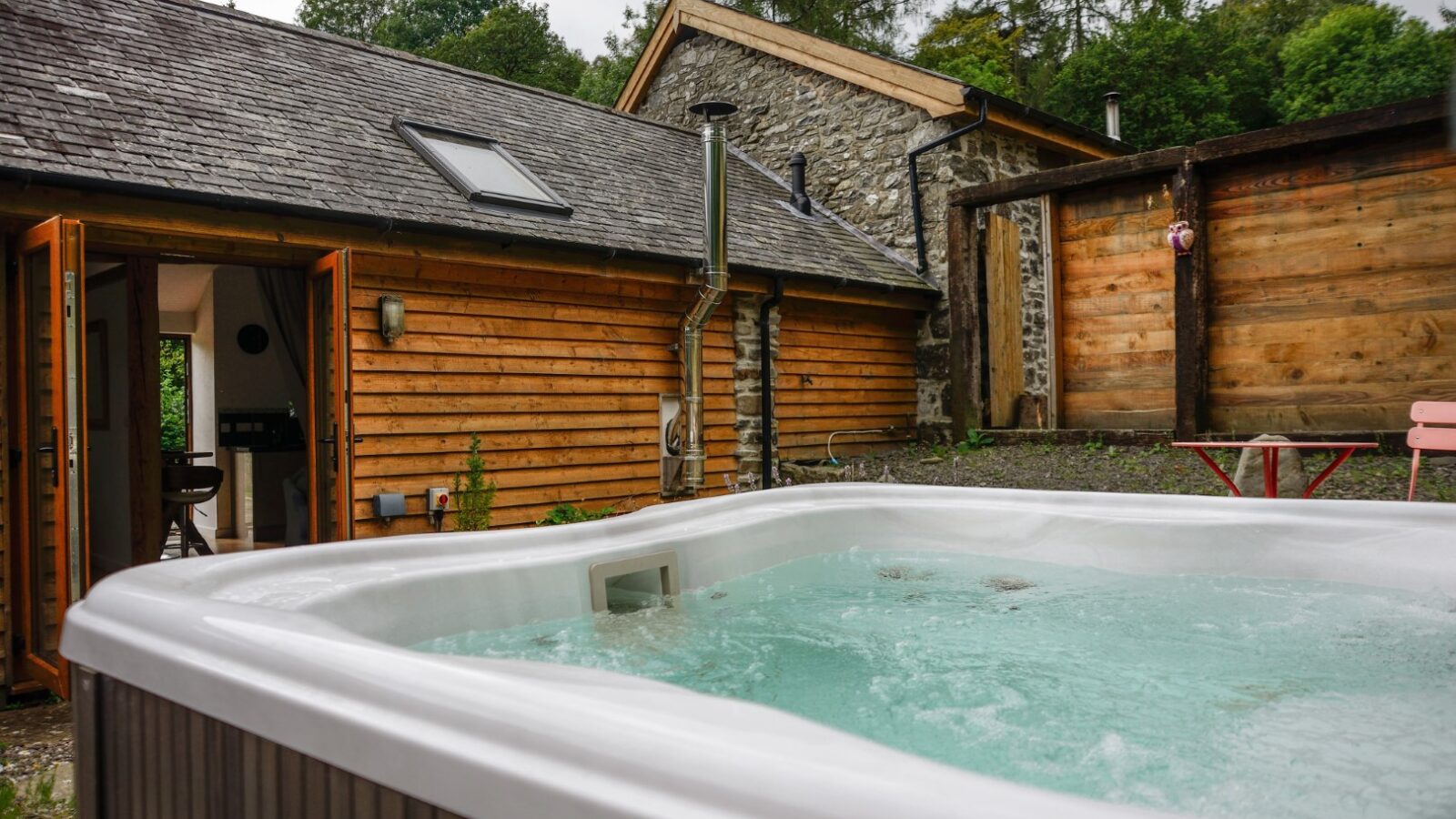 An outdoor hot tub with bubbling water sits invitingly in front of a rustic wooden and stone building with open doors at Nant Awen Cottages.