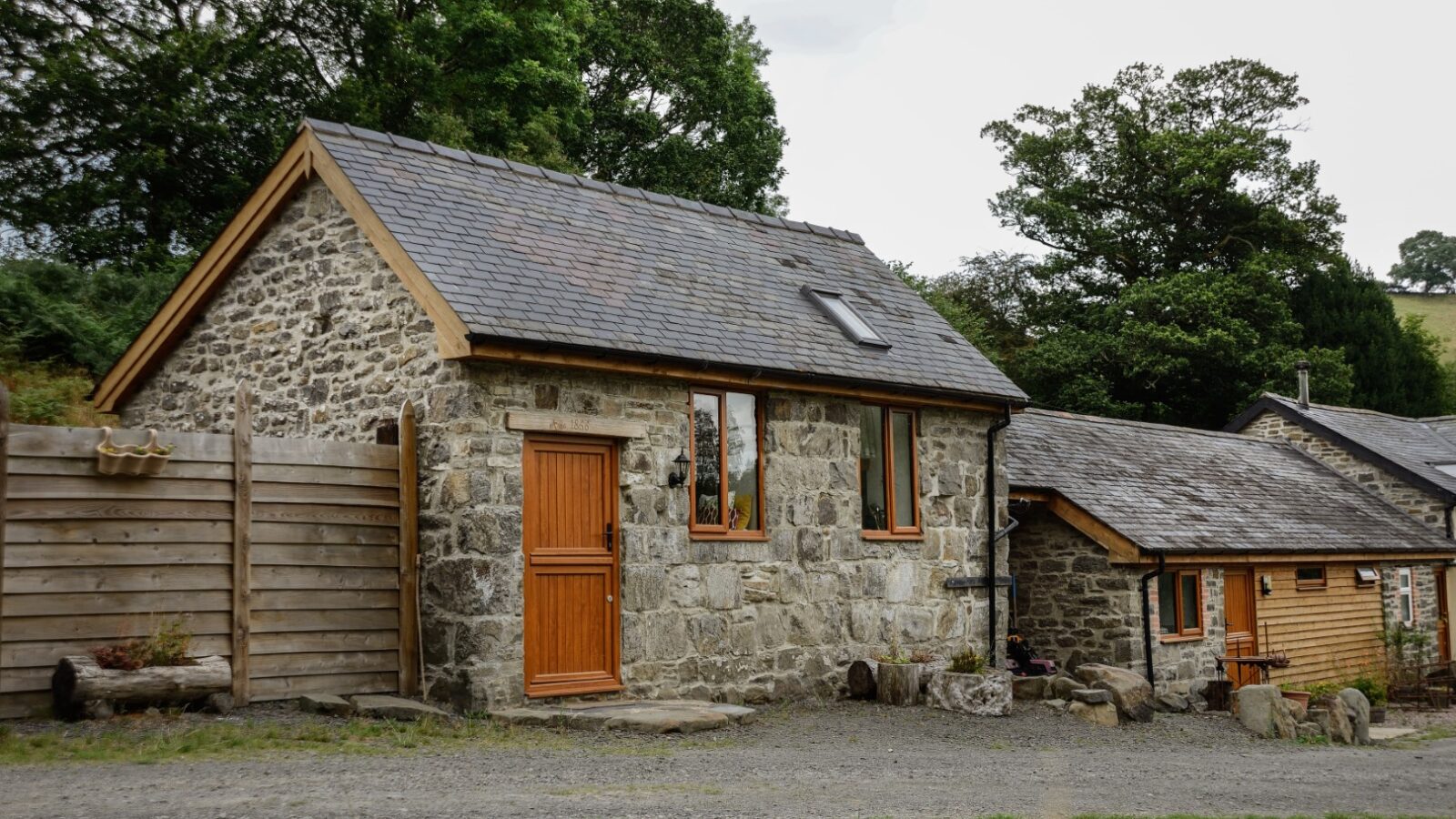 Nant Awen Cottages boasts a charming stone cottage with wooden door and window frames, nestled among trees with a peaceful dirt pathway leading through the rural landscape.