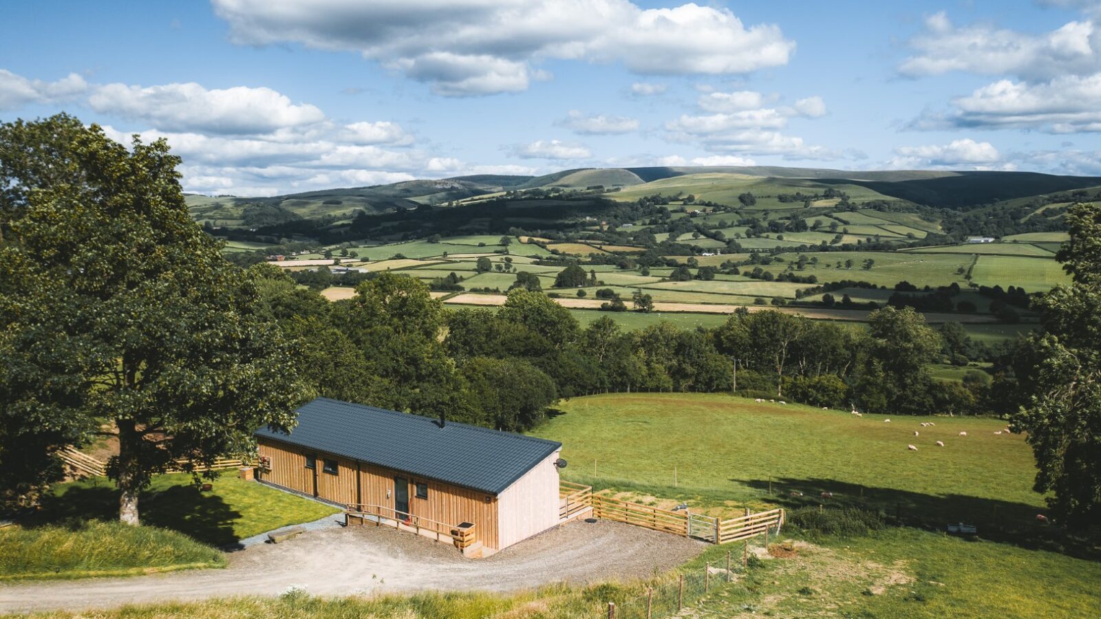 A scenic landscape featuring a charming wooden building reminiscent of Penlan lodges, surrounded by rolling hills, fields, and trees under a sky dotted with scattered clouds.