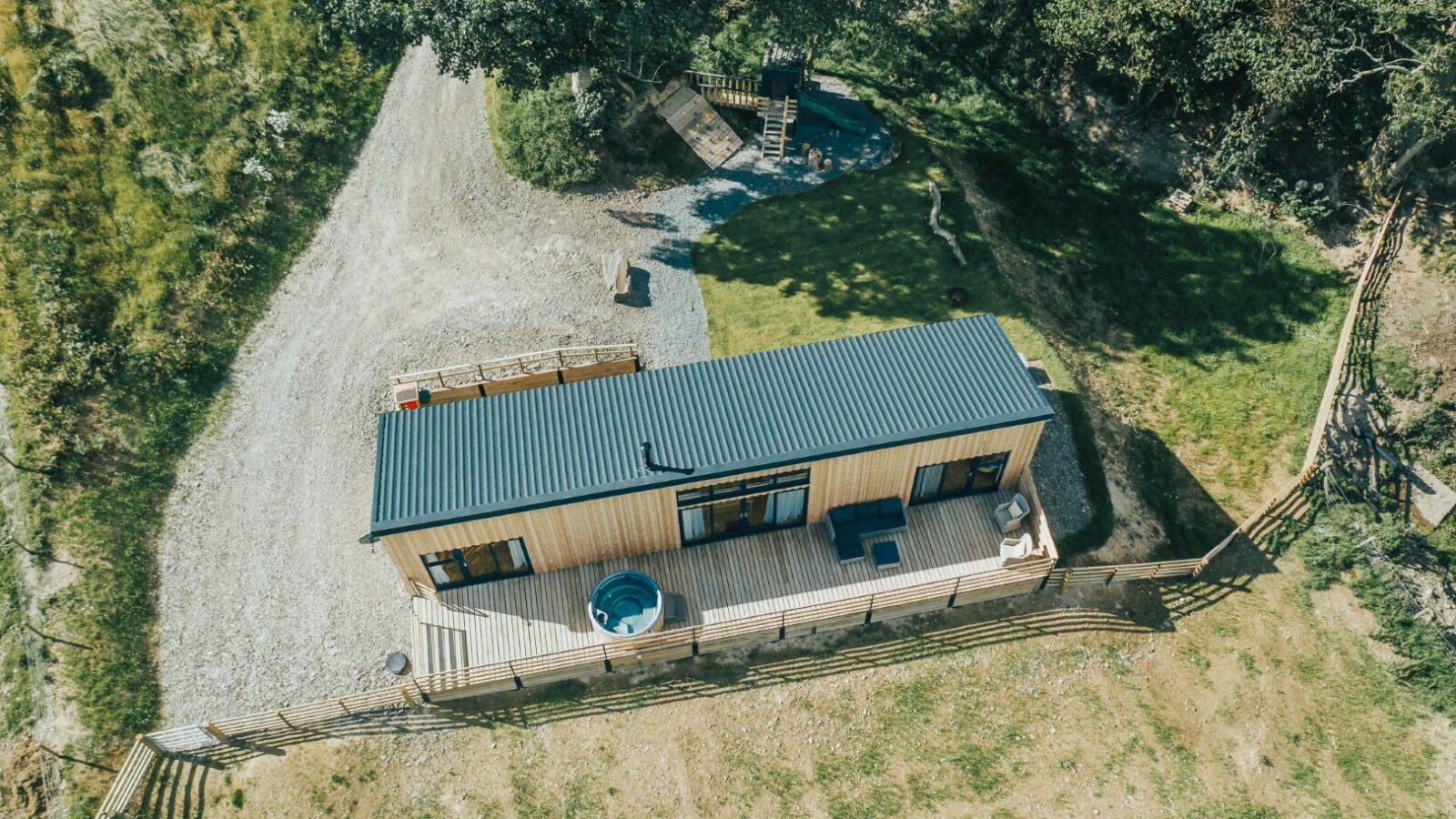 From an aerial view, the charming wooden cabin with a metal roof is nestled amidst lush grass and trees. Its inviting deck, adorned with outdoor furniture, offers a serene escape reminiscent of the tranquil ambiance found at Penlan Lodges.