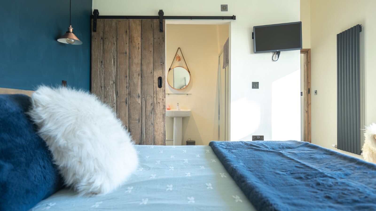 This cozy bedroom at Penlan Lodges features a striking blue accent wall, complemented by blue bedding and fluffy pillows. The room also includes a wooden sliding door and a wall-mounted TV for your entertainment.