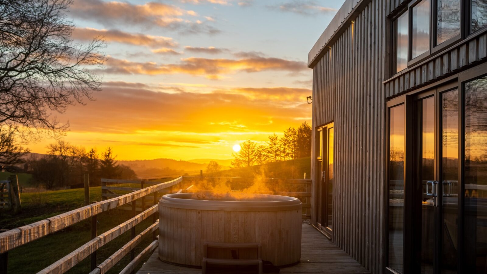 Steam rises from a hot tub on a wooden deck beside a modern building at Penlan Lodges, with a sunset and countryside view in the background.