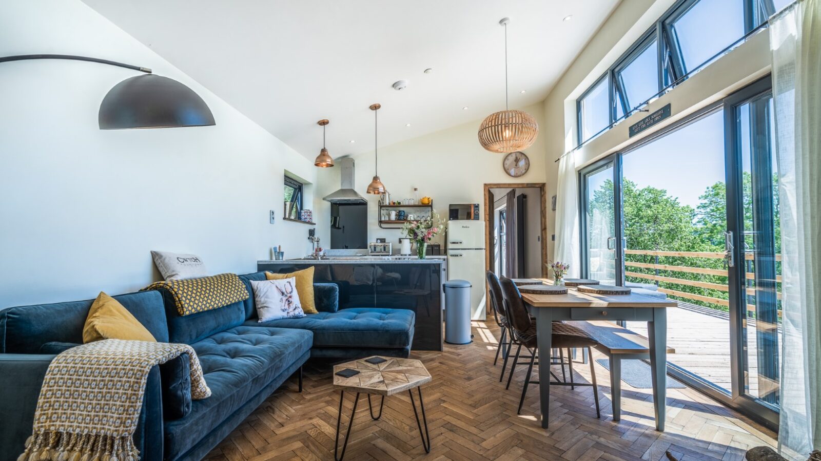 Experience the elegance of a modern open-plan living room in Penlan Lodges, featuring a velvet sofa, kitchen area, wooden dining table, and large windows opening to a balcony.