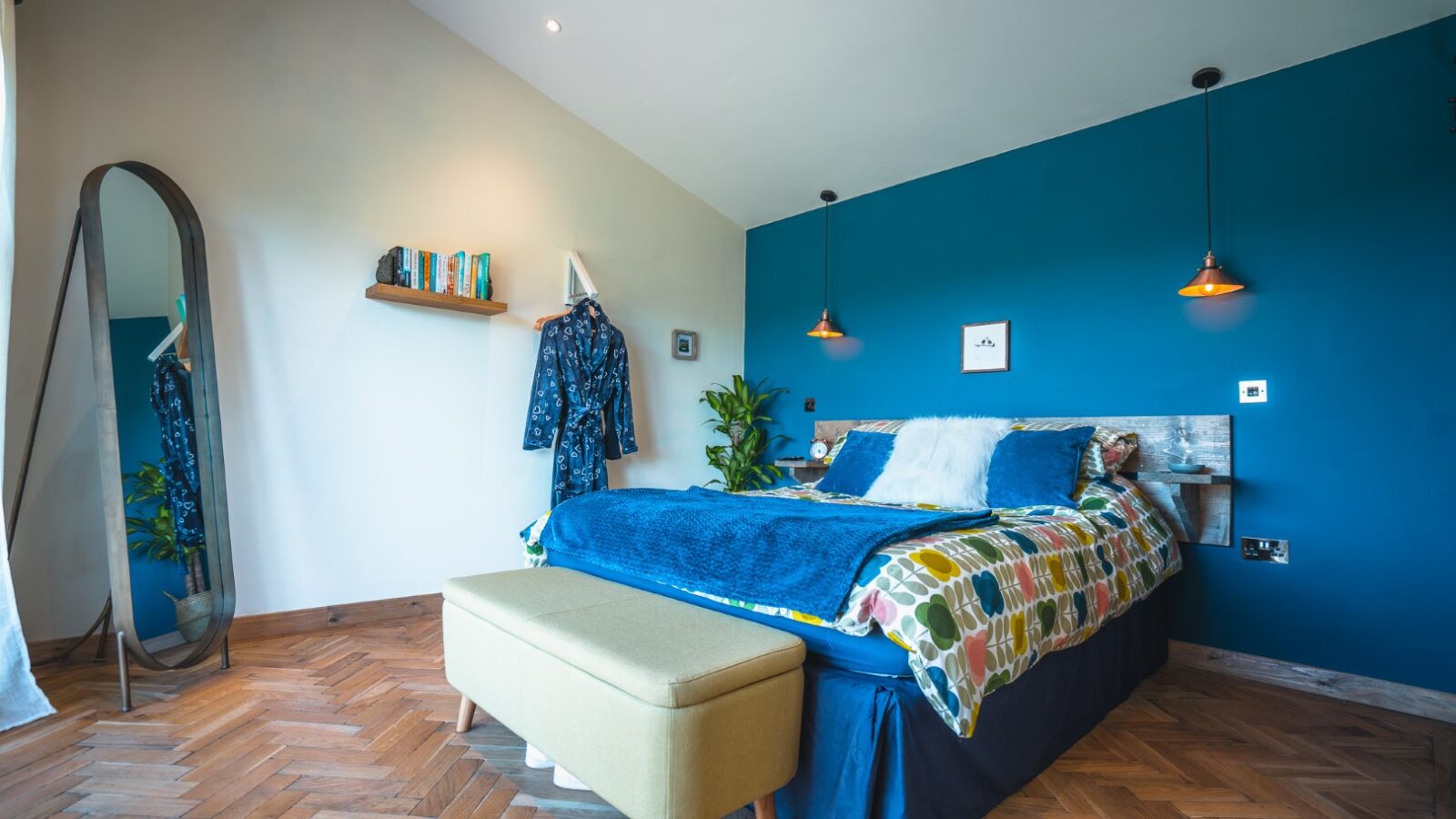 This modern bedroom at Penlan Lodges features a blue accent wall, a cozy double bed, a large mirror, lush potted plants, and a sleek bench at the foot of the bed.