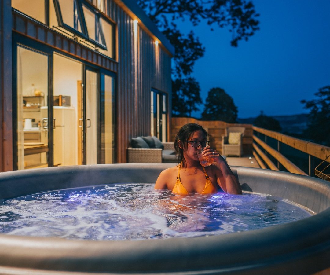 Relaxing in a hot tub at Penlan Lodges, a person enjoys a drink as the sun sets behind the cozy cabin.