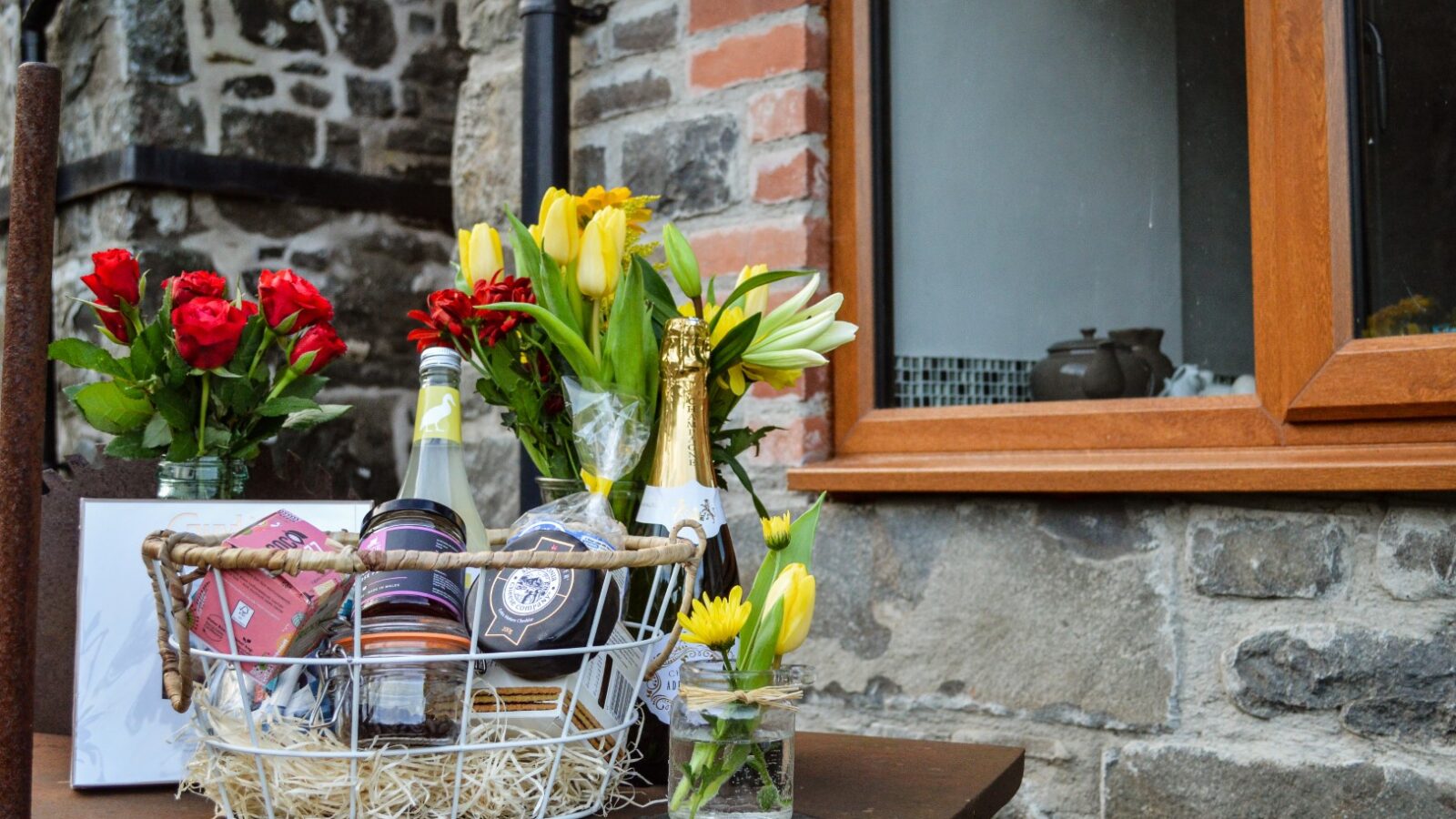 A charming gift basket with wine, chocolate, and snacks graces the table of this Nant Awen hideaway. Yellow and red flowers in vases adorn a stone wall beneath a quaint wooden window, evoking the cozy charm of traditional cottages.