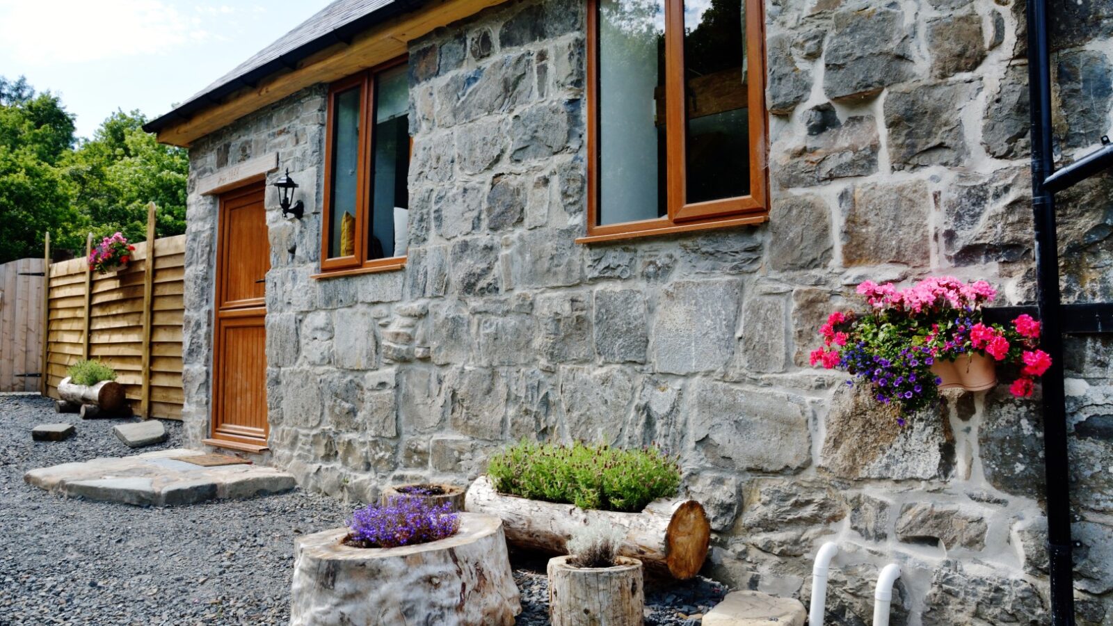 Nant Awen Cottages offers a charming stone cottage with wooden door and window frames, flower pots on logs, and pink flowers cascading down the wall, all set in a quaint gravel yard.