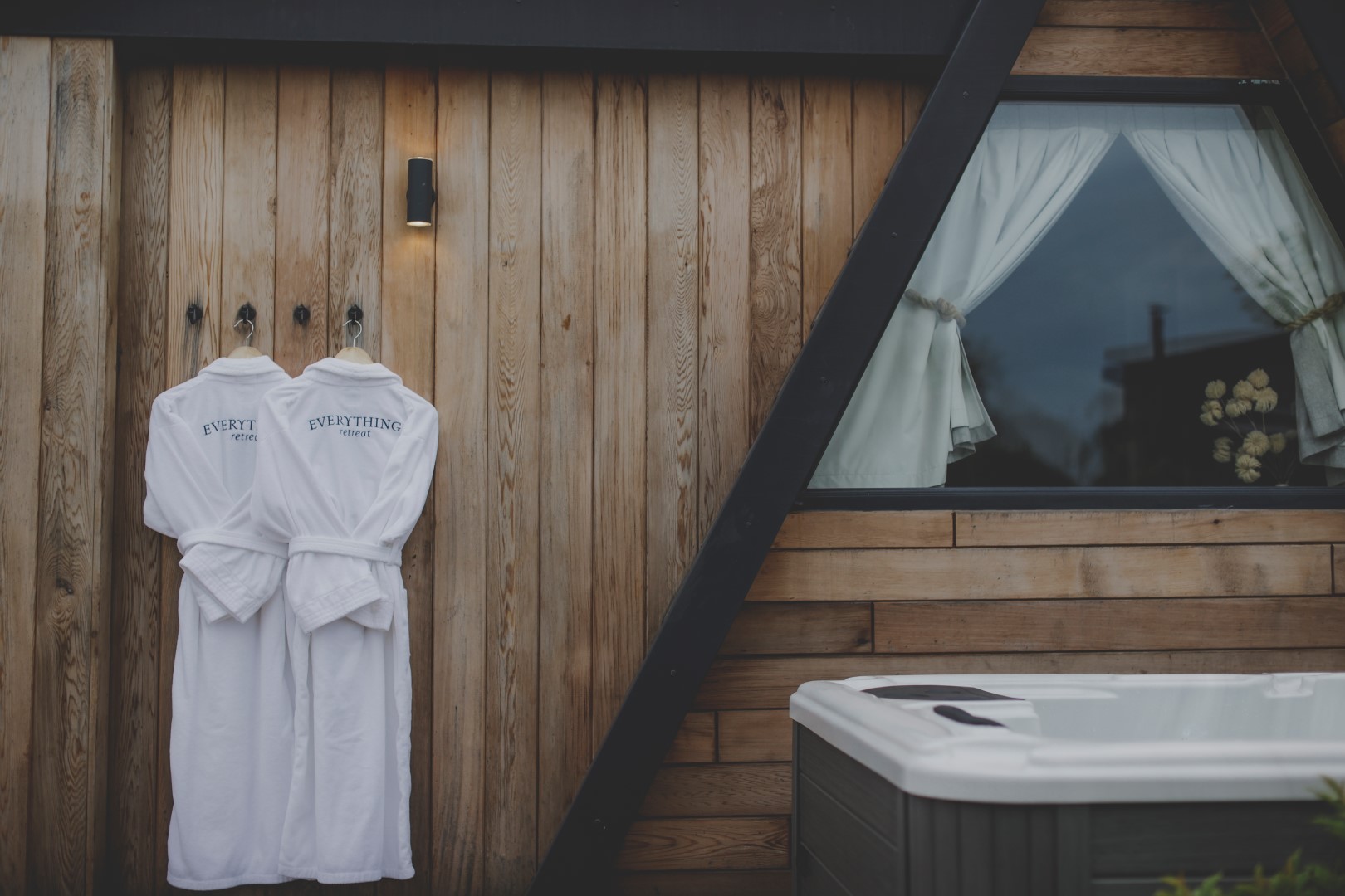 Two white bathrobes hang on hooks outside a wooden cabin with a window, epitomizing the charm of UK Hot Tub Holidays. A hot tub invites you to relax in the serene foreground.