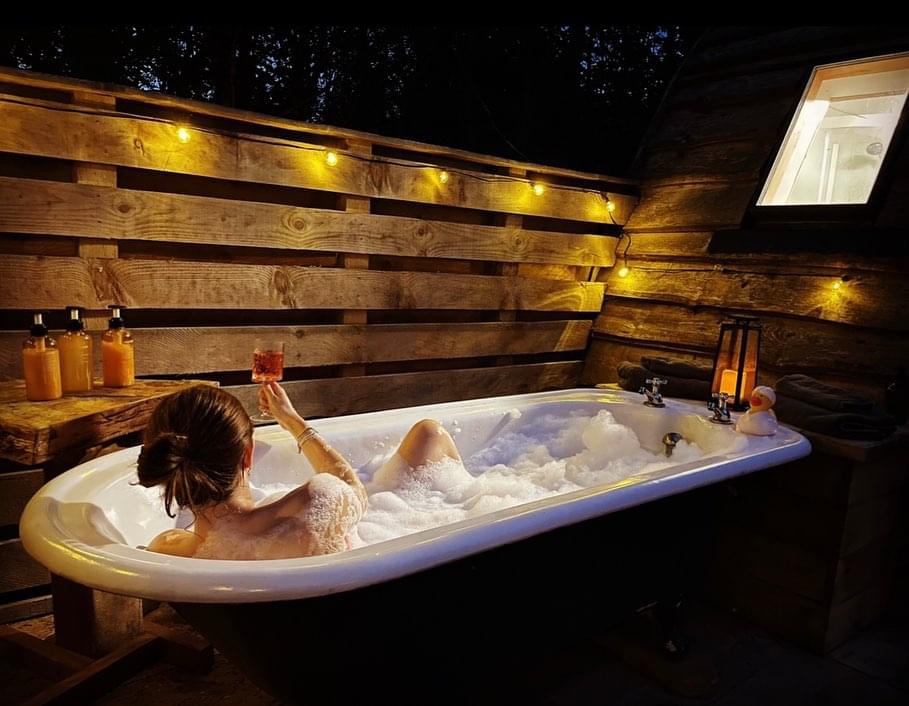 Person relaxing in an outdoor bathtub with bubbles, stargazing while holding a drink, surrounded by warm string lights and wooden decor.