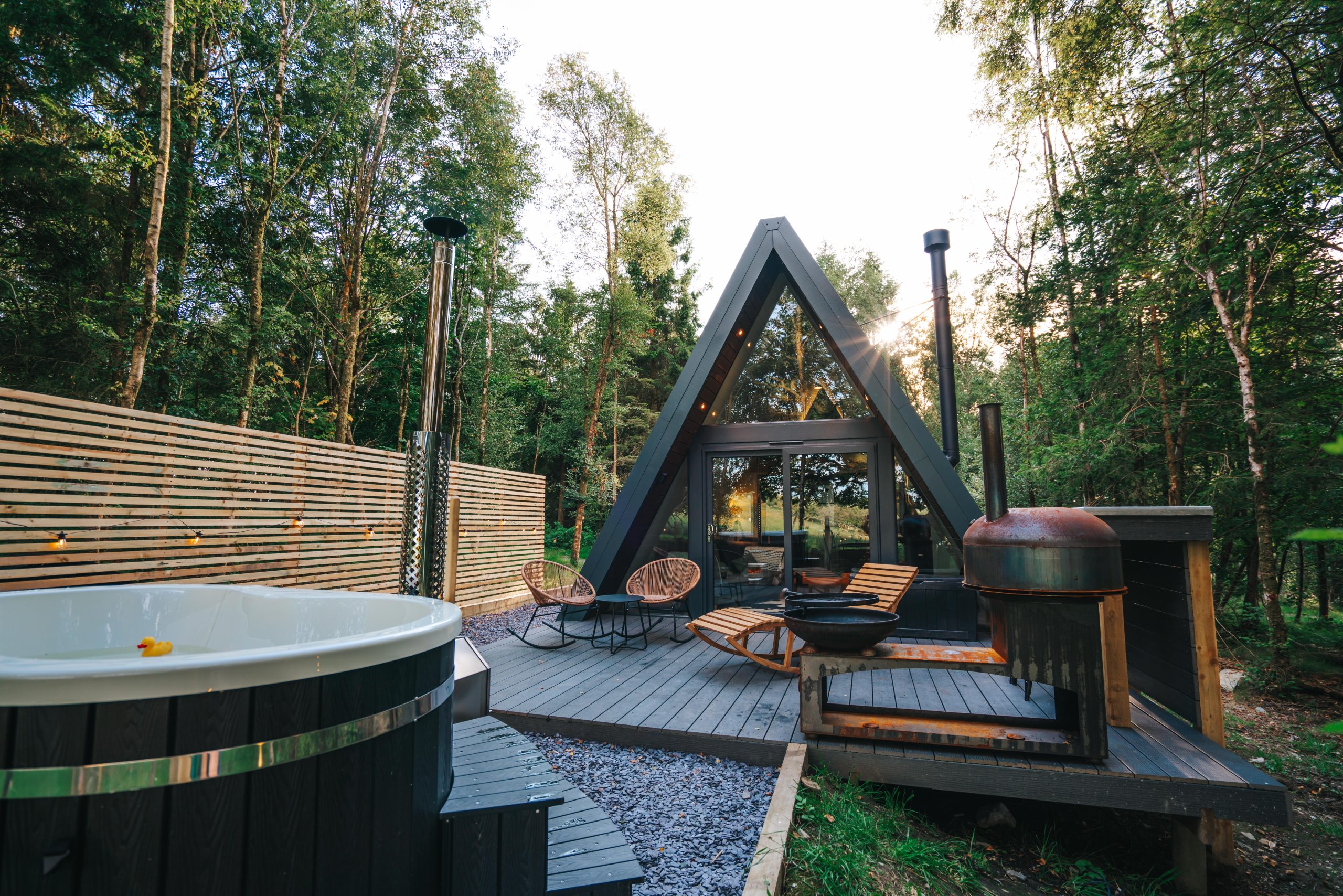 A-frame cabin with large windows, perfect for stargazing, surrounded by trees. The deck features a hot tub, lounge chairs, and a round fireplace.