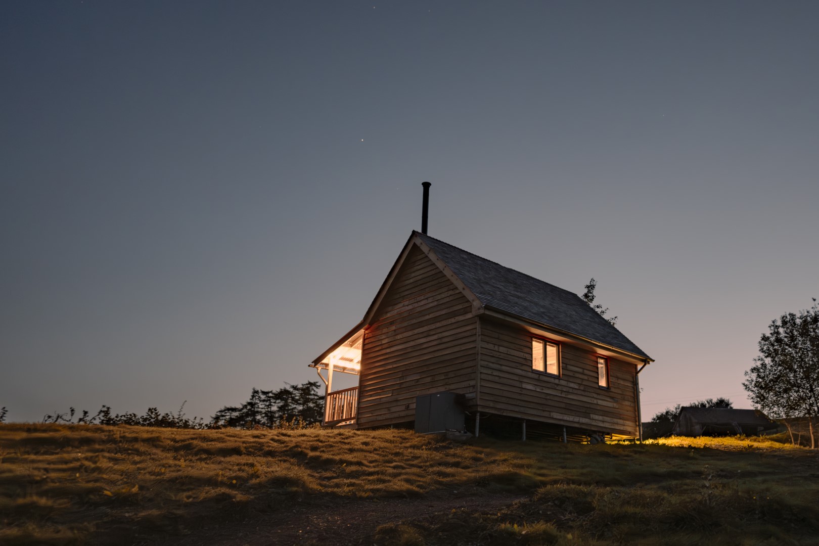 A wooden cabin perched on a grassy hill at dusk, its windows and chimney casting a warm glow, offers the perfect retreat for stargazing under a clear night sky.