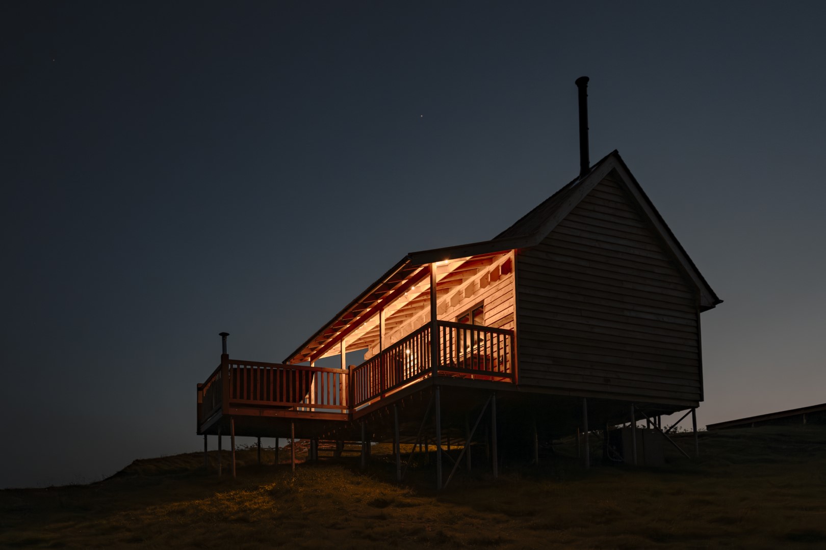 A wooden cabin on stilts with a dimly lit porch, perfect for stargazing under a dark night sky.