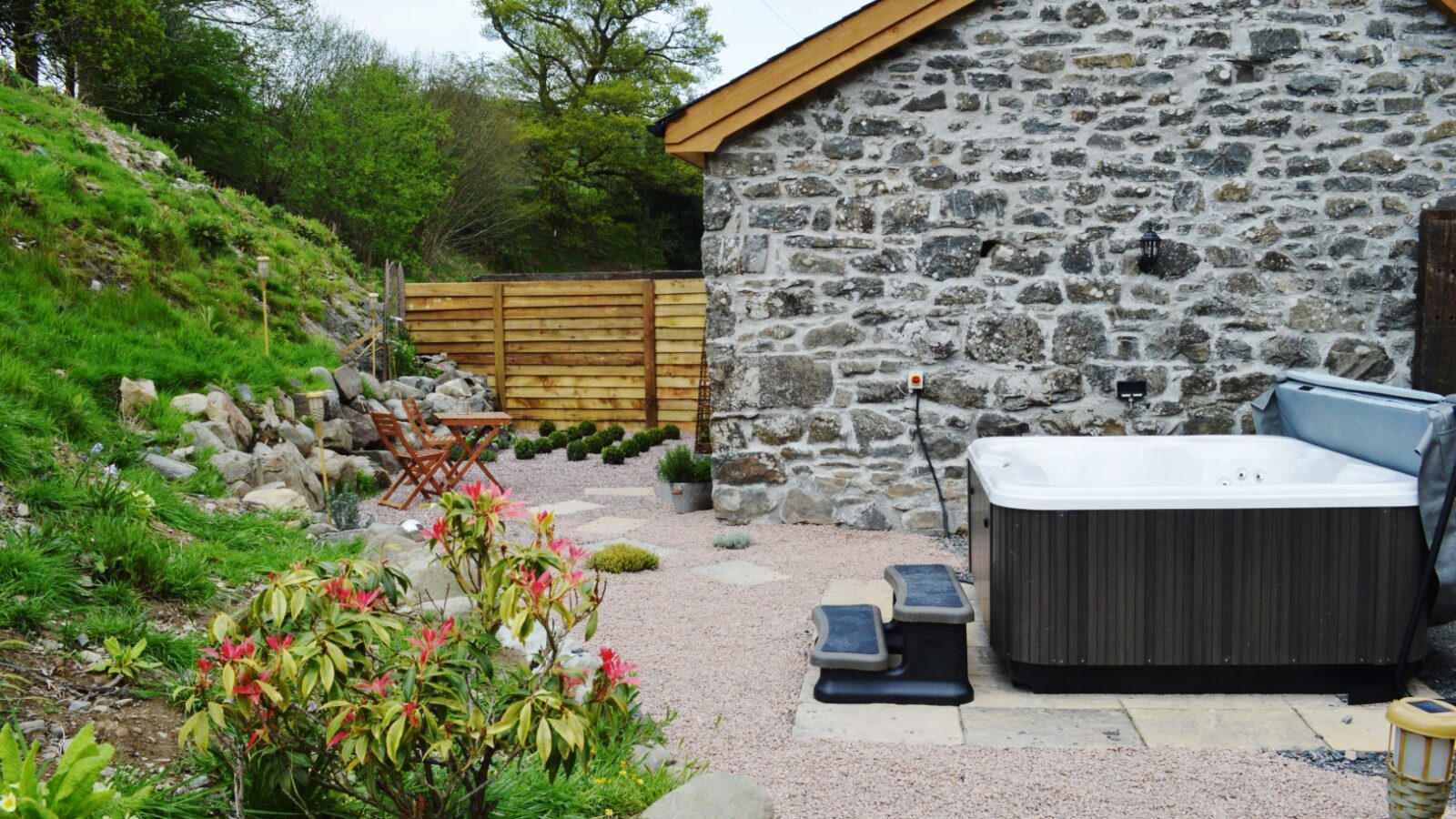 The charming stone exterior of Nant Awen Cottages features a hot tub, garden chairs, and lush plants on a gravel patio, all nestled against a verdant backdrop.