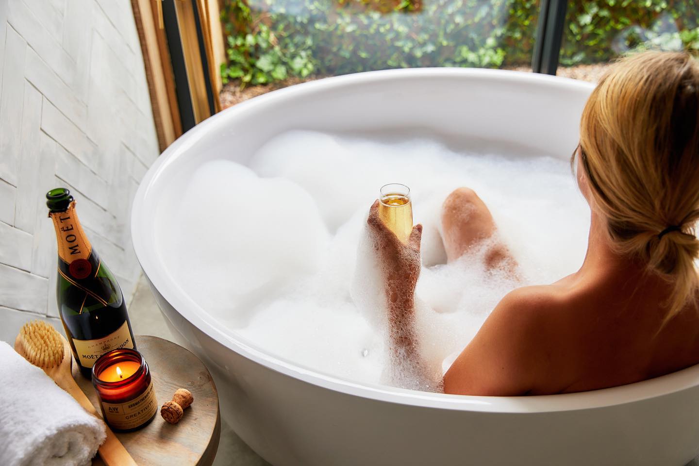 Person unwinding in a bubble bath with a glass of champagne, surrounded by the cozy scent of a candle and a bottle of Moët, embracing the essence of autumn getaways.