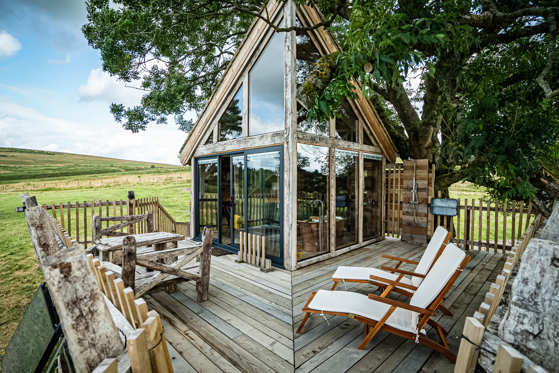 This rustic retreat treehouse boasts large windows and is nestled in a serene rural landscape, featuring a cozy deck with lounge chairs and charming outdoor seating.