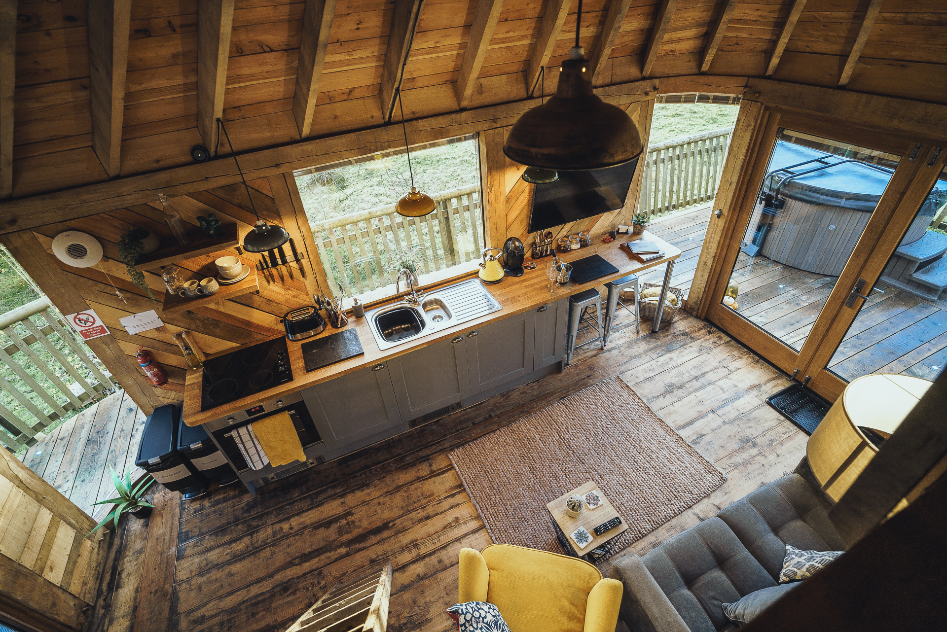 Aerial view of a cozy wooden cabin interior designed for perfect autumn getaways, with a kitchen, sofa, yellow chair, and large windows opening to a deck with a hot tub.