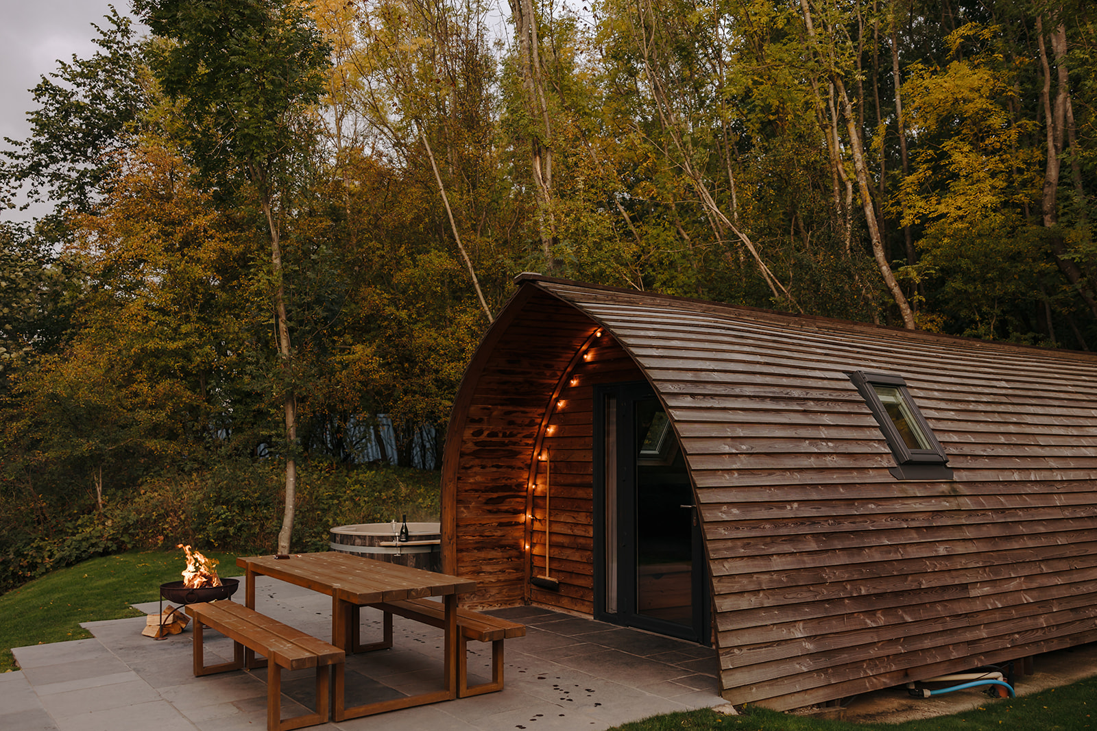 Nestled among the trees, this wooden cabin with a curved roof is the perfect spot for autumn getaways. Enjoy cozy evenings by the lit fire pit or gather around the picnic table on the patio and soak in the serene beauty of fall.