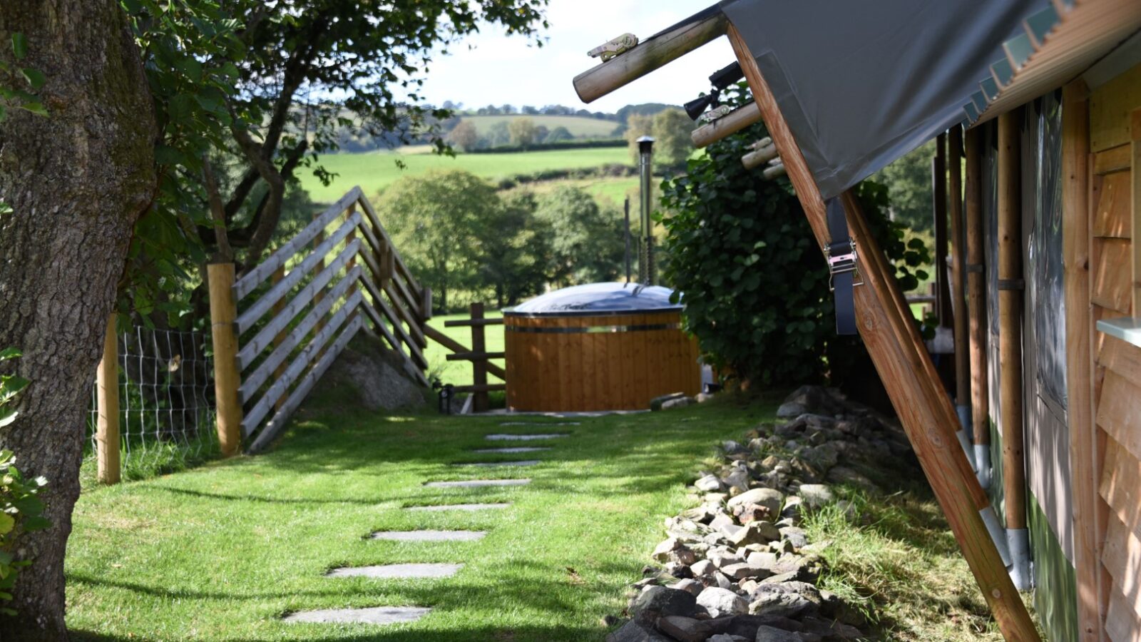 A pathway with stepping stones guides you to a wooden hot tub beside the Stellar Safari Lodge, overlooking a hilly landscape and fenced area.