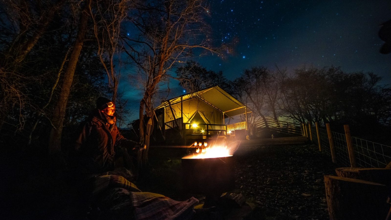 A person sits by a campfire at night, with the glowing tent of the Stellar Safari Lodge behind them, all beneath a mesmerizing starry sky. The ultimate for glamping with a hot tub in Wales