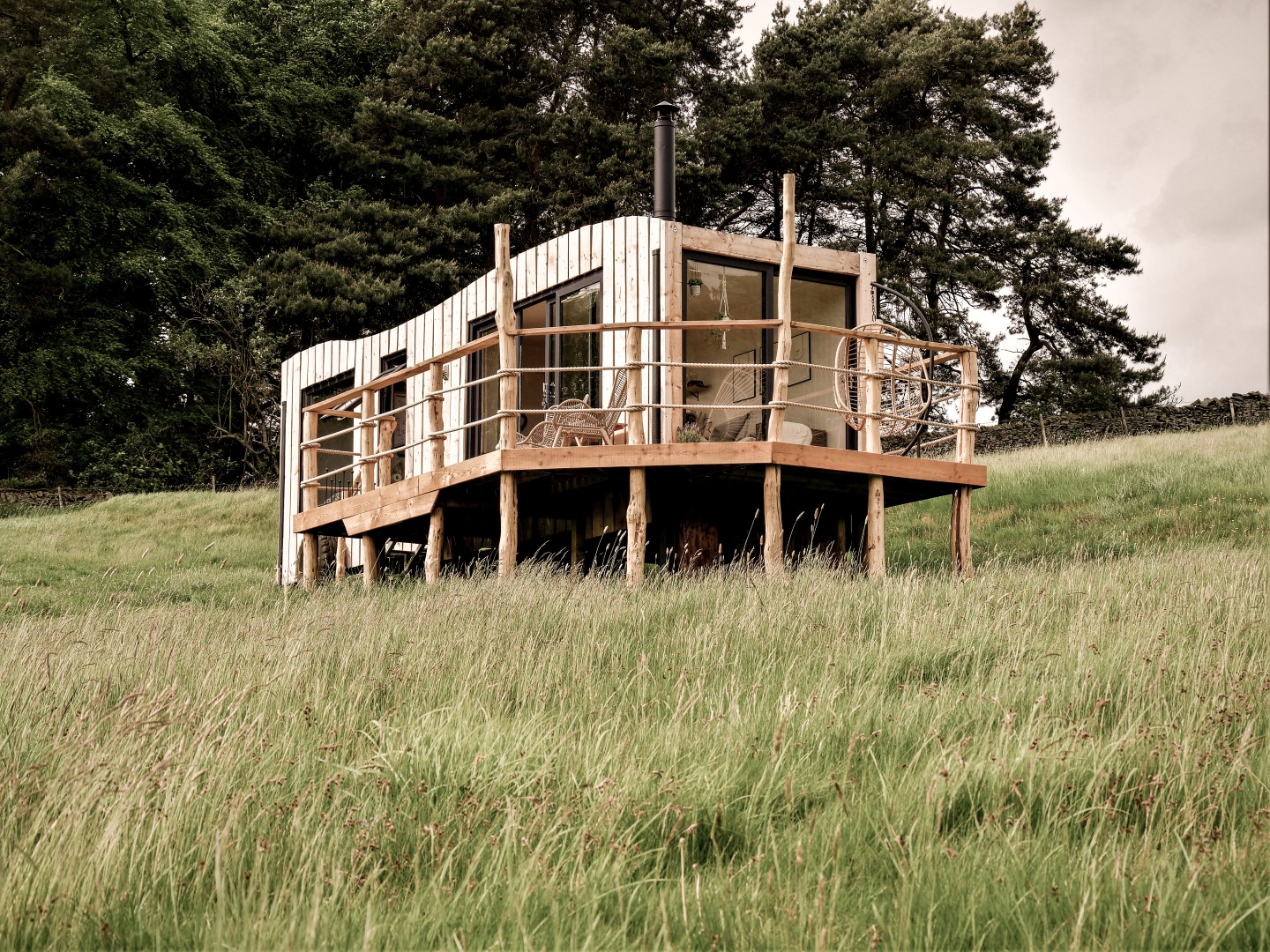 A small elevated wooden cabin, perfect for rustic retreats, is surrounded by grass and trees. It features a charming rustic railing and large windows that invite nature inside.