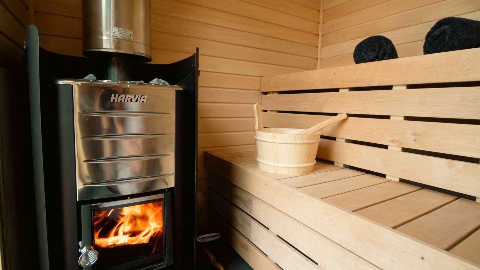 The interior of the wooden sauna at Stellar Safari Lodge features a metal stove on the left and a bucket resting on a wooden bench to the right, offering an authentic rustic retreat amidst nature.