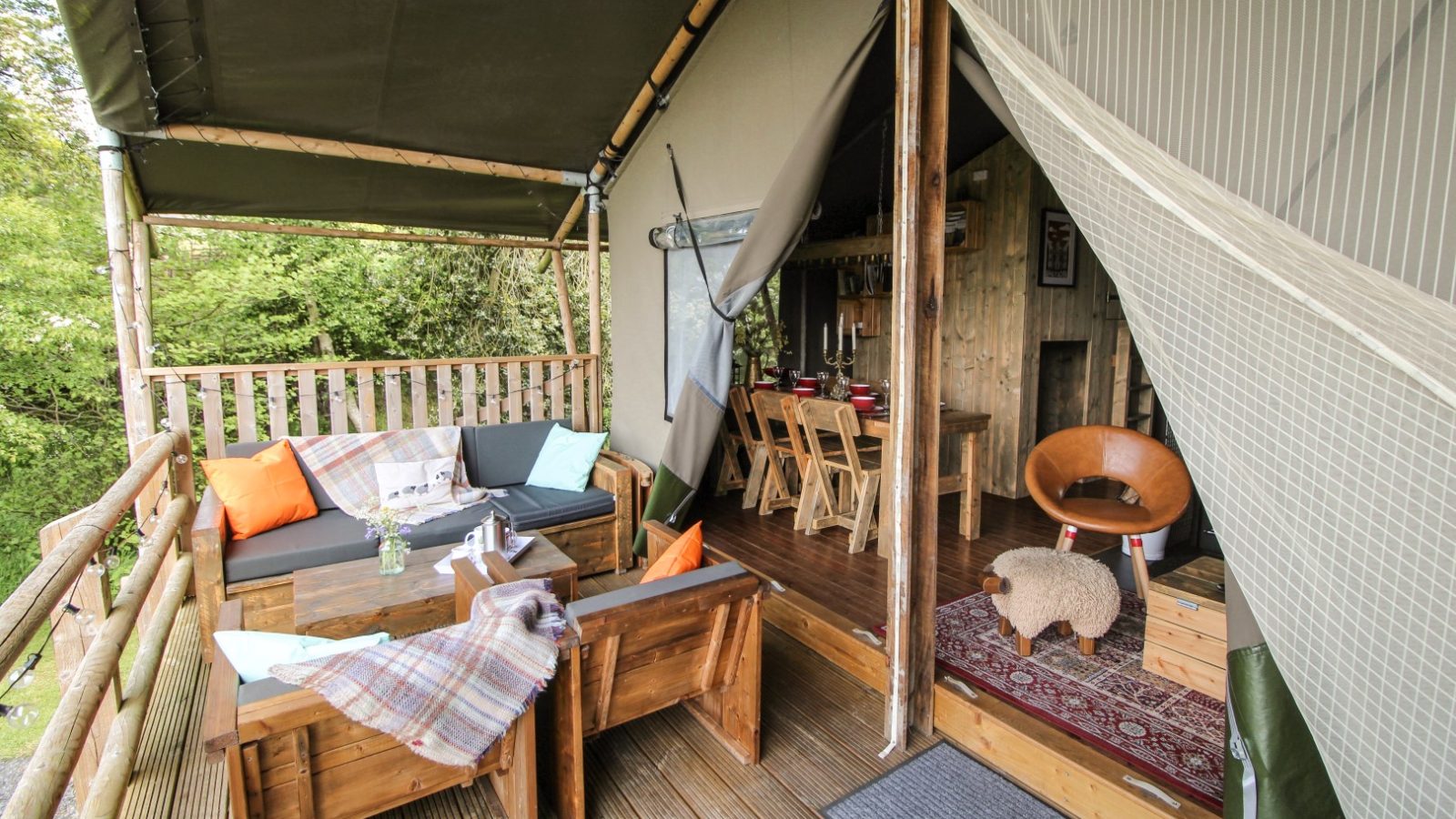 The Stellar Safari Lodge offers an eco-friendly cabin interior featuring wooden furniture and colorful cushions in the dining area, all surrounded by lush greenery.