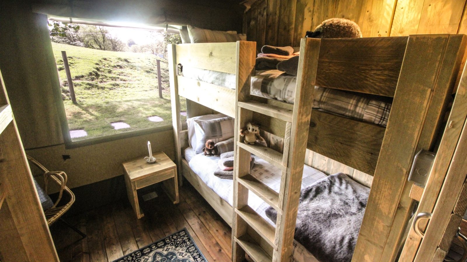 The bunk bed in the rustic wooden room offers a true Stellar Safari Lodge experience, complete with a window view of a grassy field. A small table and chair are thoughtfully placed beside it, perfect for soaking in the tranquil ambiance.