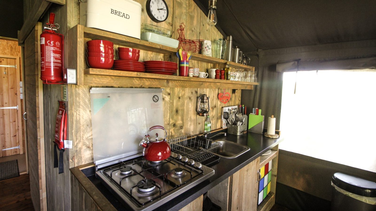 A rustic kitchen reminiscent of a Stellar Safari Lodge, featuring wooden shelves, a vibrant red kettle on the stove, stacked red dishes, and a charming wall clock. Sunlight streams through the window, enhancing the lodge-like ambiance.