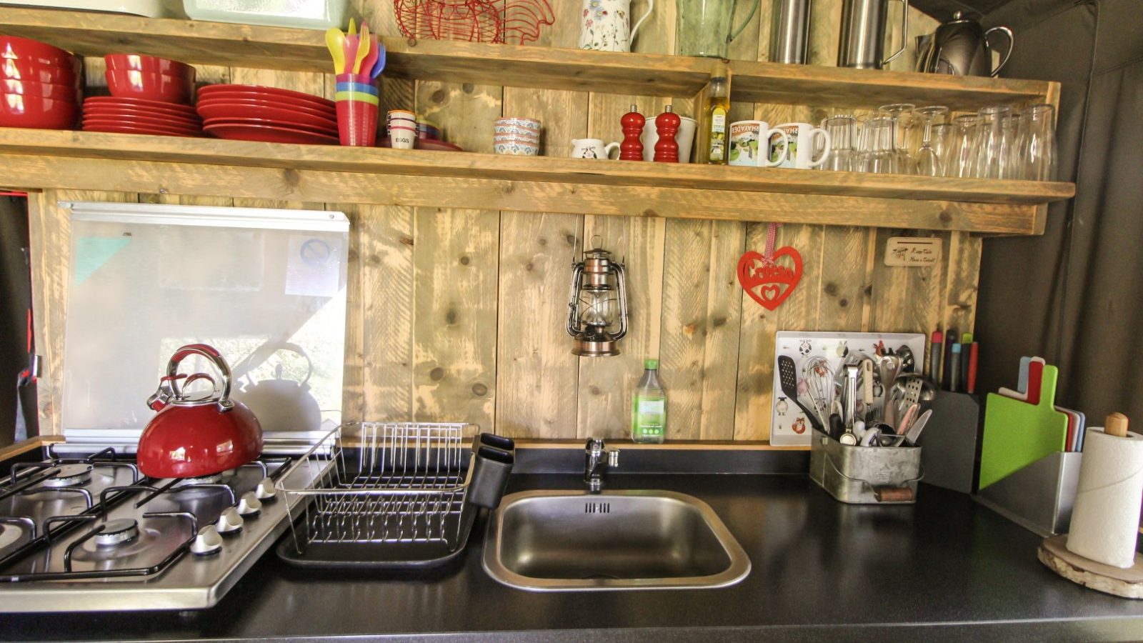 The Stellar Safari Lodge-themed wooden kitchen features a stove, red kettle, dish rack, and utensils. Rustic shelves hold red dishes, cups, glasses, and various kitchen items that evoke the charm of an adventurous escape.