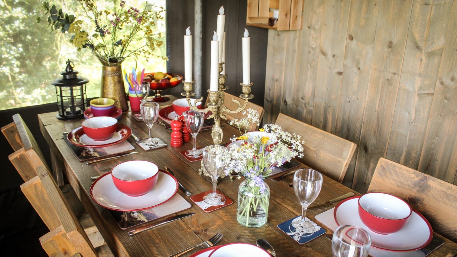 A wooden dining table at the Stellar Safari Lodge is set with vibrant red dishware, elegant wine glasses, a classic candelabra, and fresh flowers in jars.