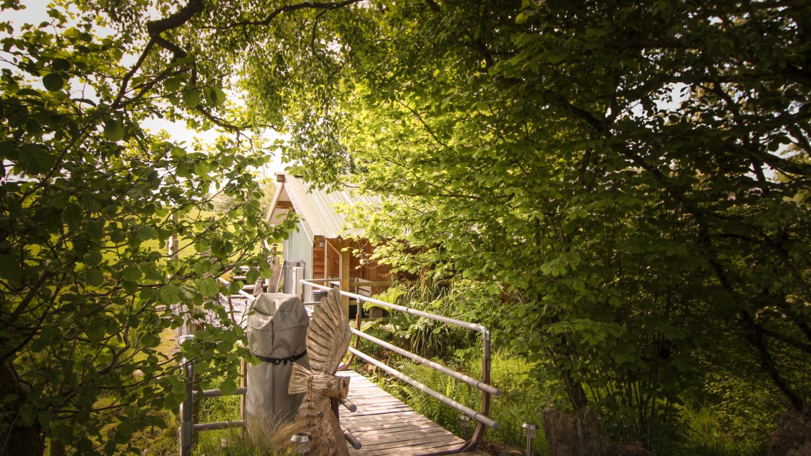 The wooden bridge guides you to the Stellar Safari Lodge, a charming treehouse nestled among lush green foliage on a sunny day.