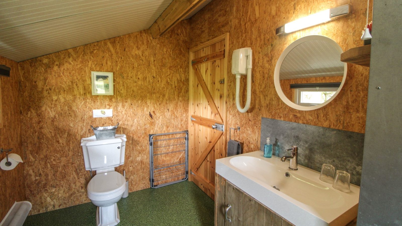 Experience the charm of a Stellar Safari Lodge in this bathroom, featuring wood-paneled walls, a white toilet, round mirror, sink with toiletries, and an overhead light.