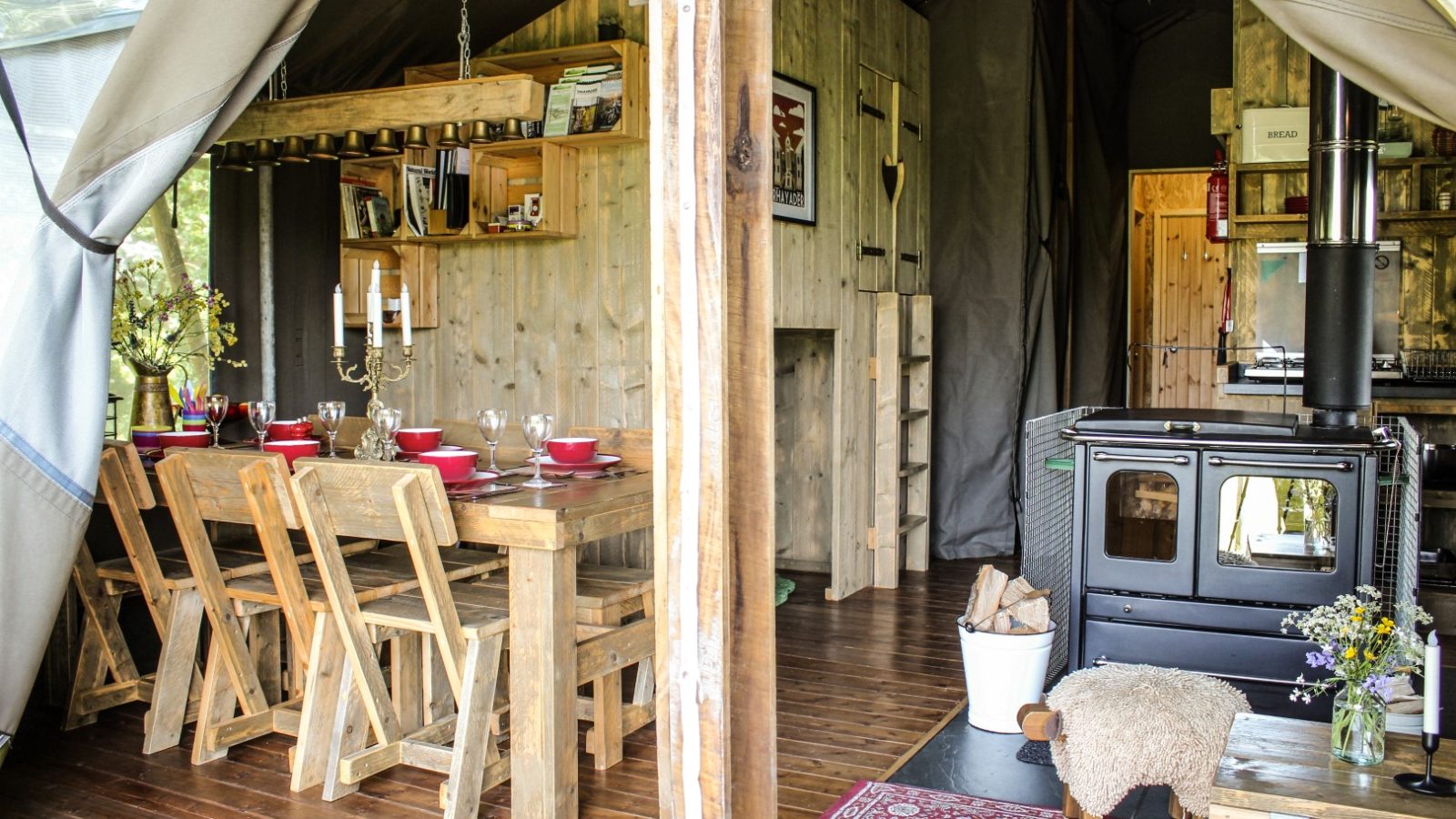 Rustic dining and living area in a wooden cabin at Stellar Safari Lodge, featuring a stove, dining table, chairs, and shelves adorned with various items.