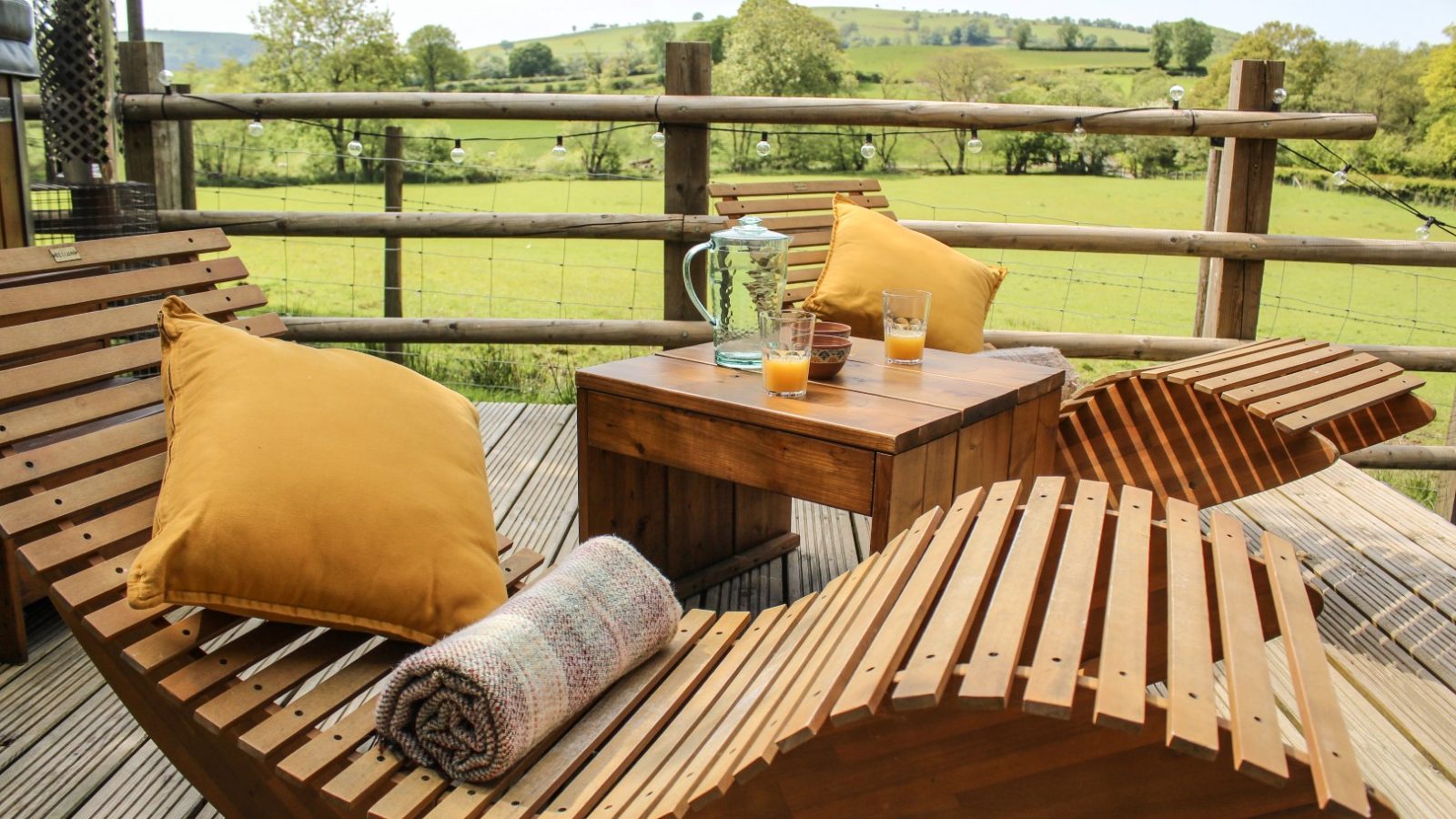 The wooden deck at Stellar Safari Lodge features two lounge chairs with yellow pillows, a small table adorned with drinks, and offers a stunning view of the green field and rolling hills.