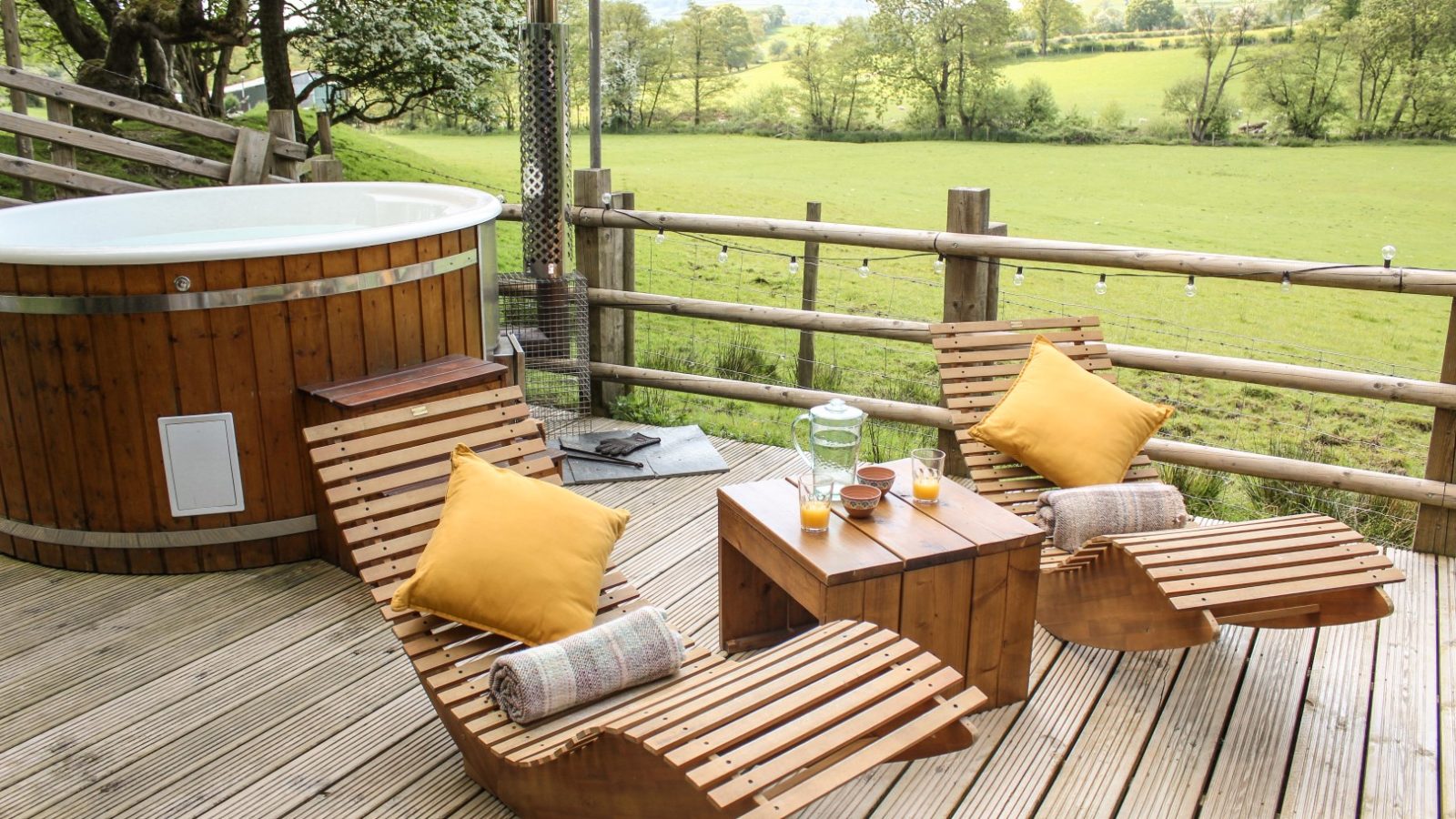 At the Stellar Safari Lodge, two wooden lounge chairs with yellow pillows invite relaxation next to a small table on the deck. Nearby, a round hot tub offers serene views of a grassy field, perfect for unwinding amidst nature's tranquility.