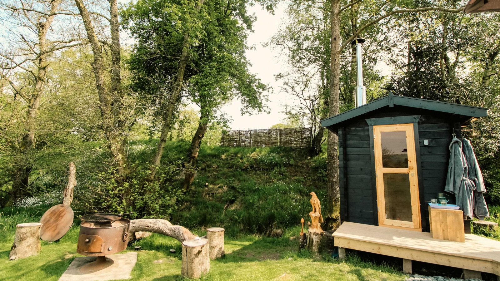 Nestled in a natural setting, the Stellar Safari Lodge features a charming wooden sauna with a chimney. Nearby, guests can relax by the fire pit on rustic tree stumps, enjoying the serene ambiance of this unique lodge experience.