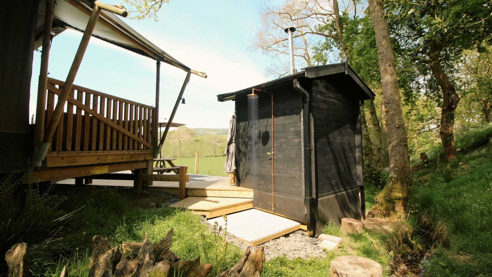 A stellar black outdoor sauna rests beside a wooden deck and tent, evoking a safari lodge ambiance amid lush trees and grass under a sunny sky.