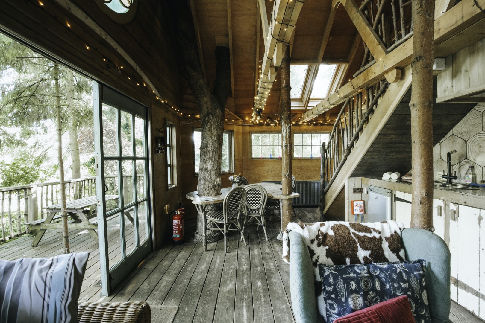 Cozy treehouse interior with wooden decor, a seating area, dining table, and stairs. Large windows offer a perfect view of the trees outside, ideal for autumn getaways.