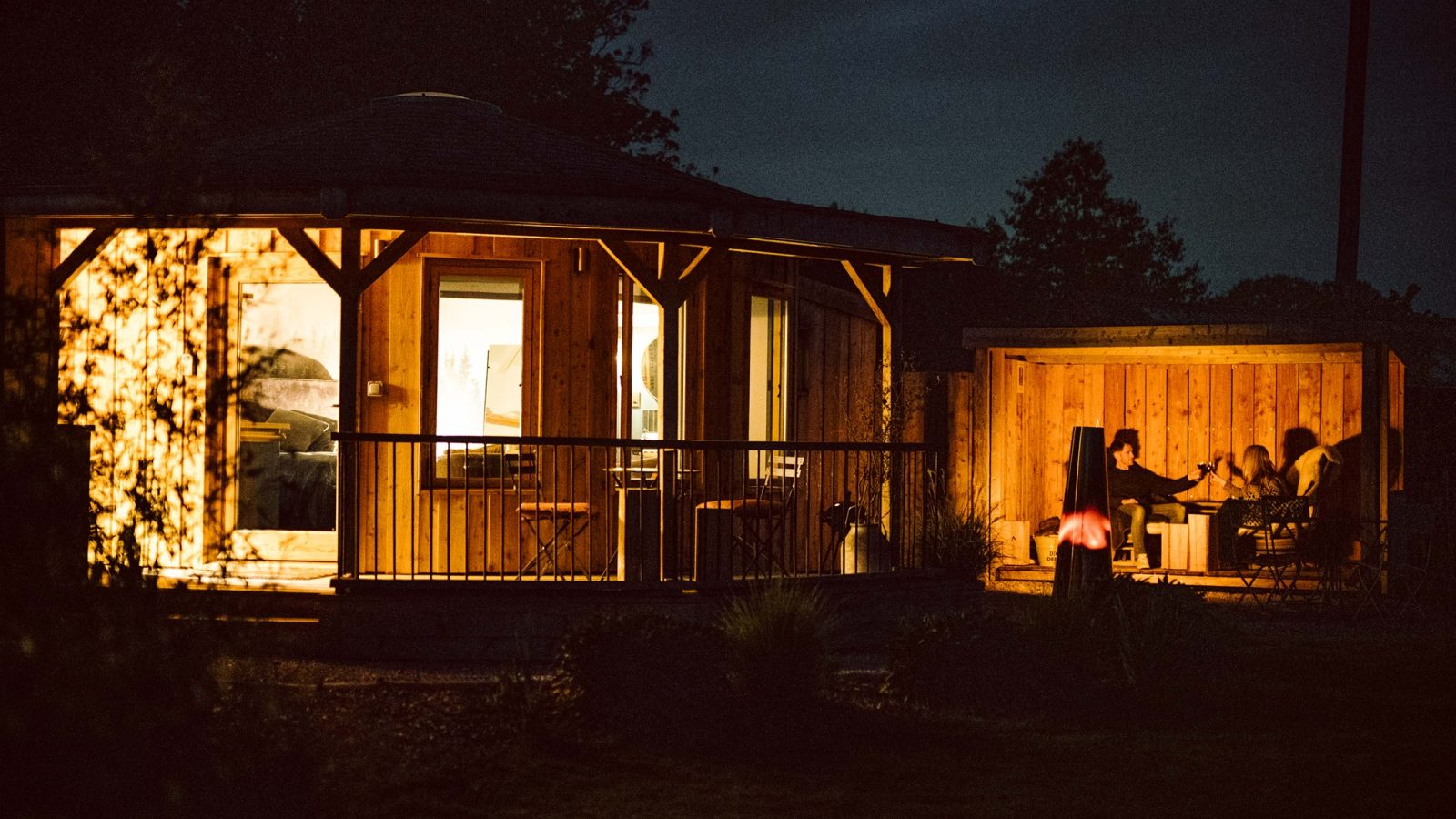 An inviting outdoor seating area at night, enhanced by warm lighting, features people gathered around a table. In the background, a round wooden building adds charm, making it perfect for cozy autumn getaways.