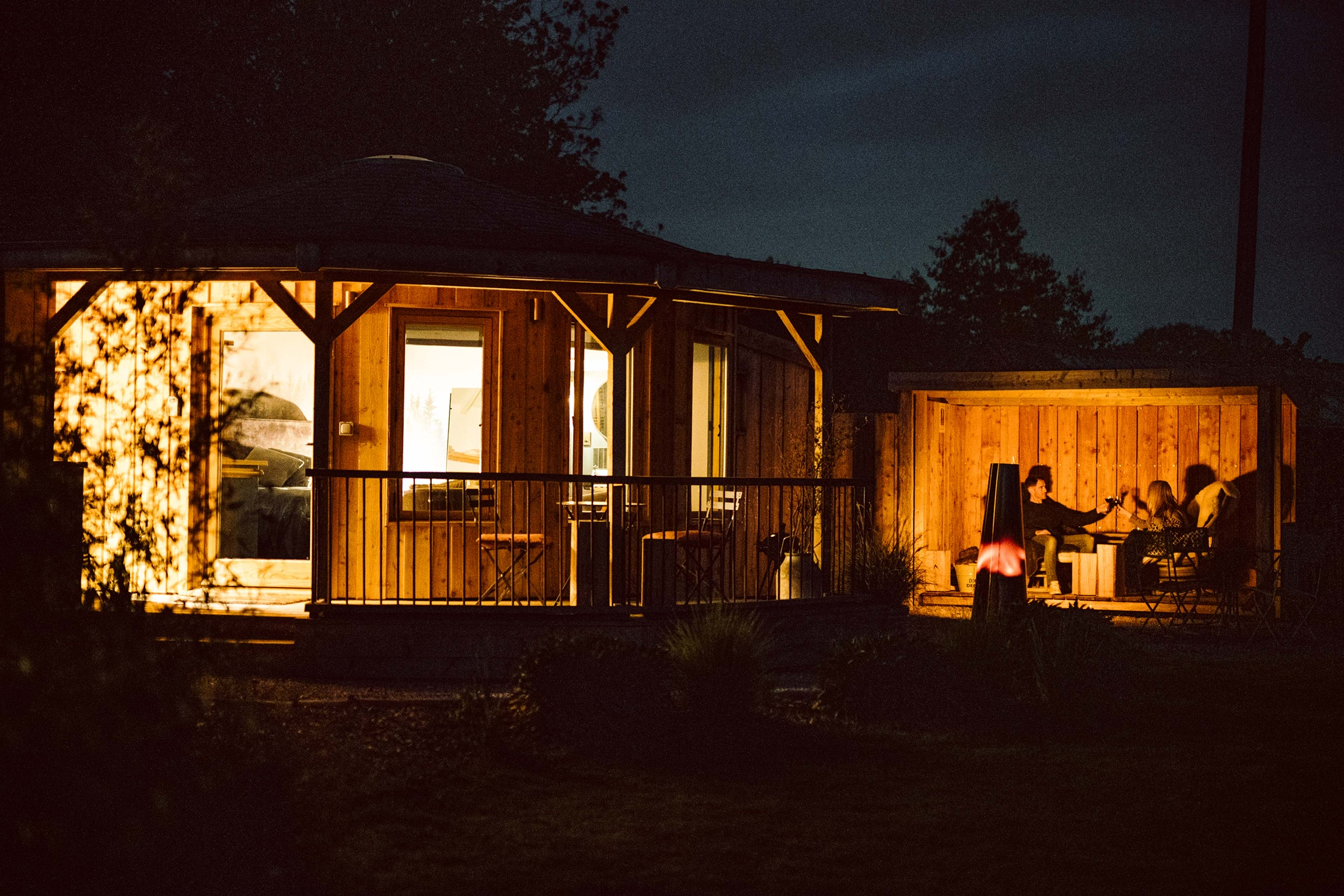 An inviting outdoor seating area at night, enhanced by warm lighting, features people gathered around a table. In the background, a round wooden building adds charm, making it perfect for cozy autumn getaways.