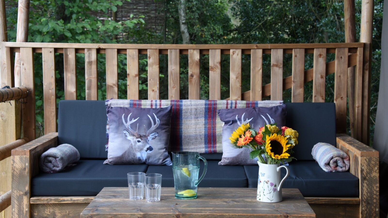 Outdoor patio with a wooden sofa, deer-themed cushions, and Safari-inspired vibes. The table is set with a clear pitcher and glasses next to a floral jug brimming with sunflowers, reminiscent of a cozy lodge feel.