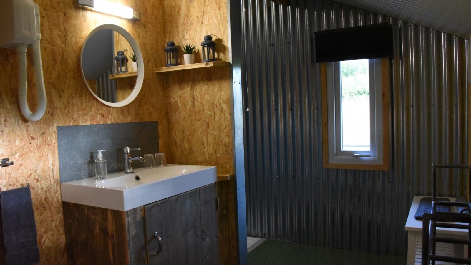 A rustic bathroom at the Stellar Safari Lodge features a wooden vanity, wall-mounted round mirror, and corrugated metal walls, providing an authentic Safari experience.