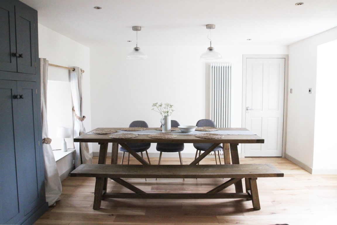 A minimalist dining area features a wooden table, bench, and chairs. Embracing the charm of autumn getaways, two pendant lights hang overhead, casting a warm glow on the vase centerpiece.