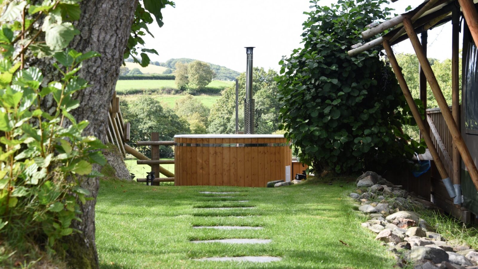 A stone path leads to a wooden hot tub amid lush greenery and trees, creating a serene retreat reminiscent of a stellar safari lodge nestled in nature.